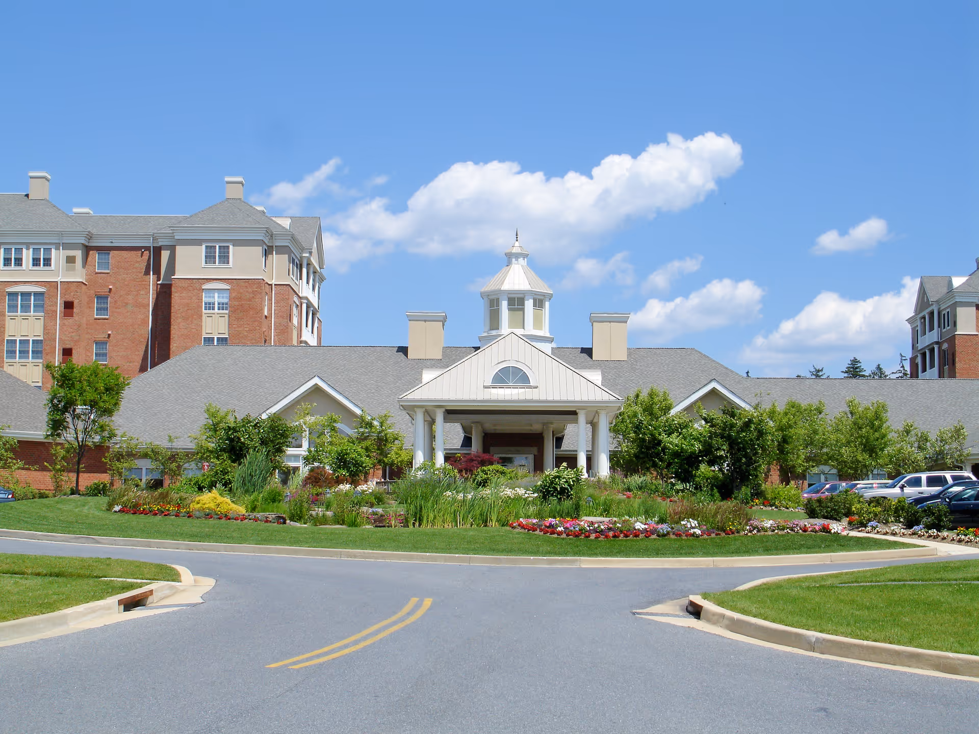 Front exterior view of Mercy Ridge facility showing a large building with a central entrance featuring white columns and a cupola on the roof. The building is surrounded by well-maintained landscaping with green shrubs, colorful flowers, and trees under a blue sky with scattered clouds. There is a paved driveway leading up to the entrance.
