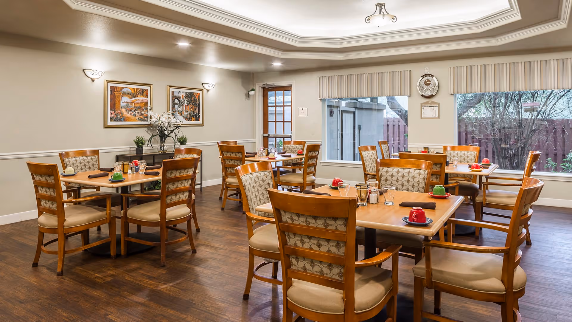 A dining room with several wooden tables and chairs arranged neatly. Each table has colorful cups and glasses set on it. The room has large windows with striped valances, allowing natural light to enter. The walls are decorated with framed artwork and sconces, and there is a wooden floor.