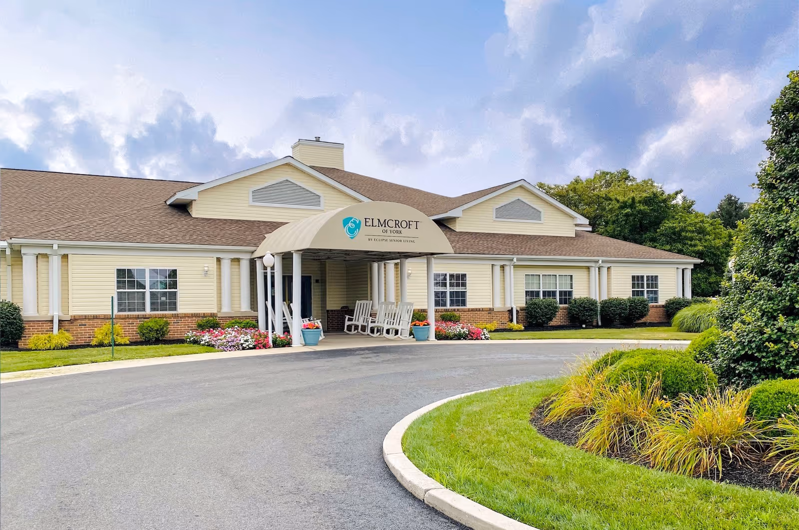 Exterior view of a single-story senior living facility building with a covered entrance featuring rocking chairs. The building has beige siding with brick accents and is surrounded by well-maintained landscaping including green bushes, colorful flowers, and a curved driveway. The sky is partly cloudy.