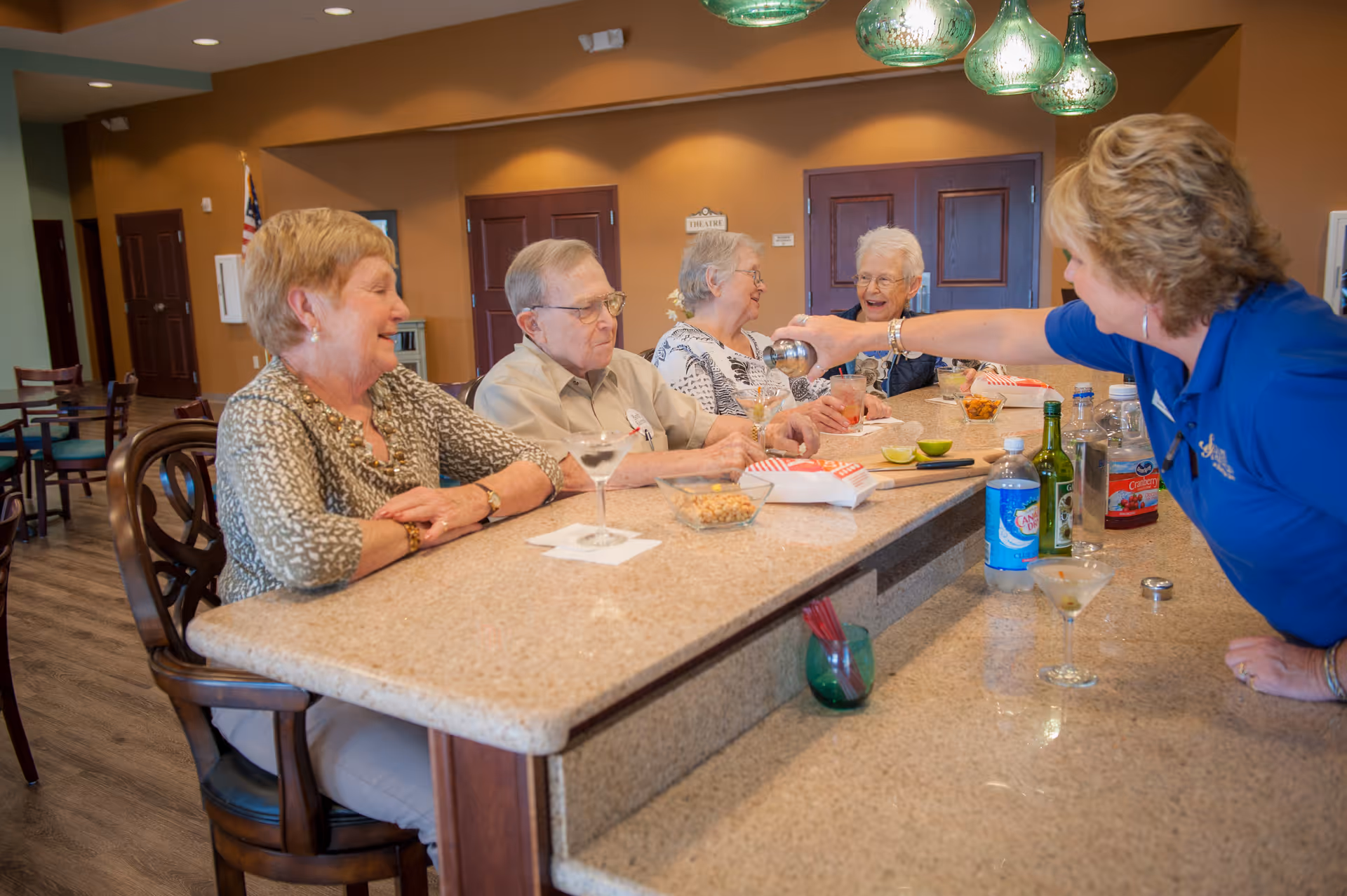 Four elderly residents sit at a community bar counter while a staff member serves drinks and snacks in a bright common dining area.