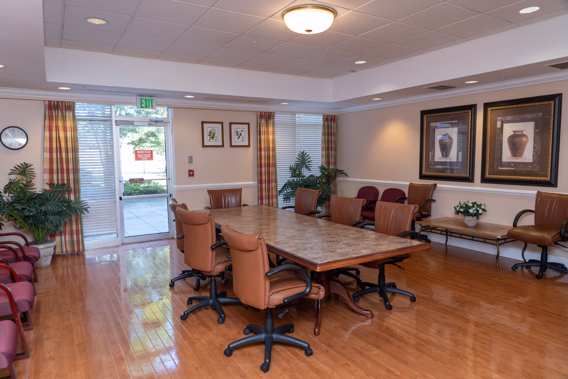 A well-lit meeting or dining room with a large rectangular table surrounded by brown leather swivel chairs. The room has wooden flooring, framed artwork on the walls, potted plants, and windows with plaid curtains. There is a glass door leading outside with an exit sign above it.