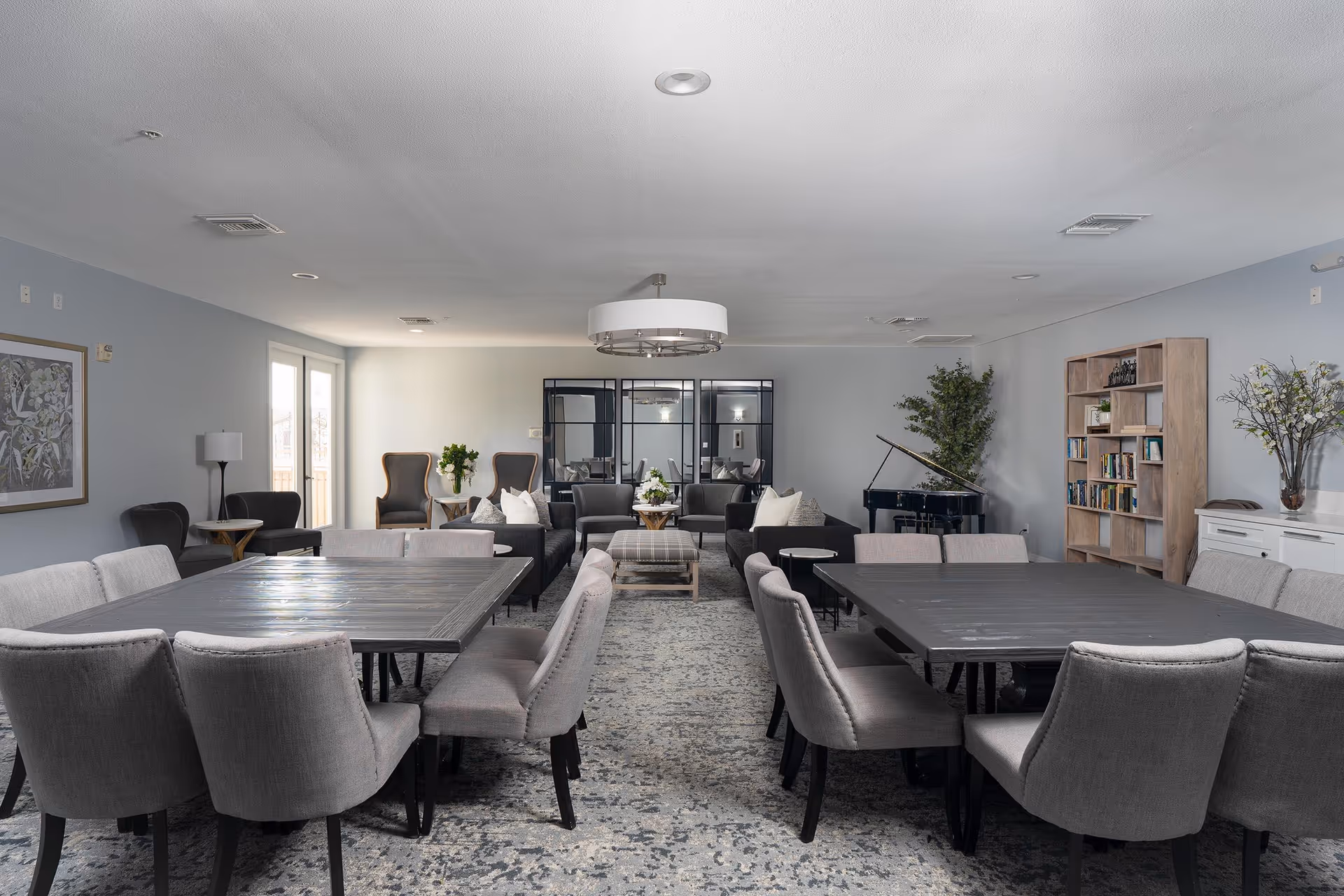 A spacious and well-lit common area featuring two large dark wooden tables surrounded by gray upholstered chairs. In the background, there are several black armchairs, a black grand piano, a wooden bookshelf filled with books, and decorative plants. The room has light gray walls, a patterned carpet, and a modern ceiling light fixture.