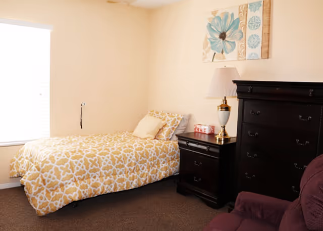 A small furnished bedroom with a single bed in a yellow patterned comforter, a dark wood nightstand and tall dresser, table lamp, and wall art.