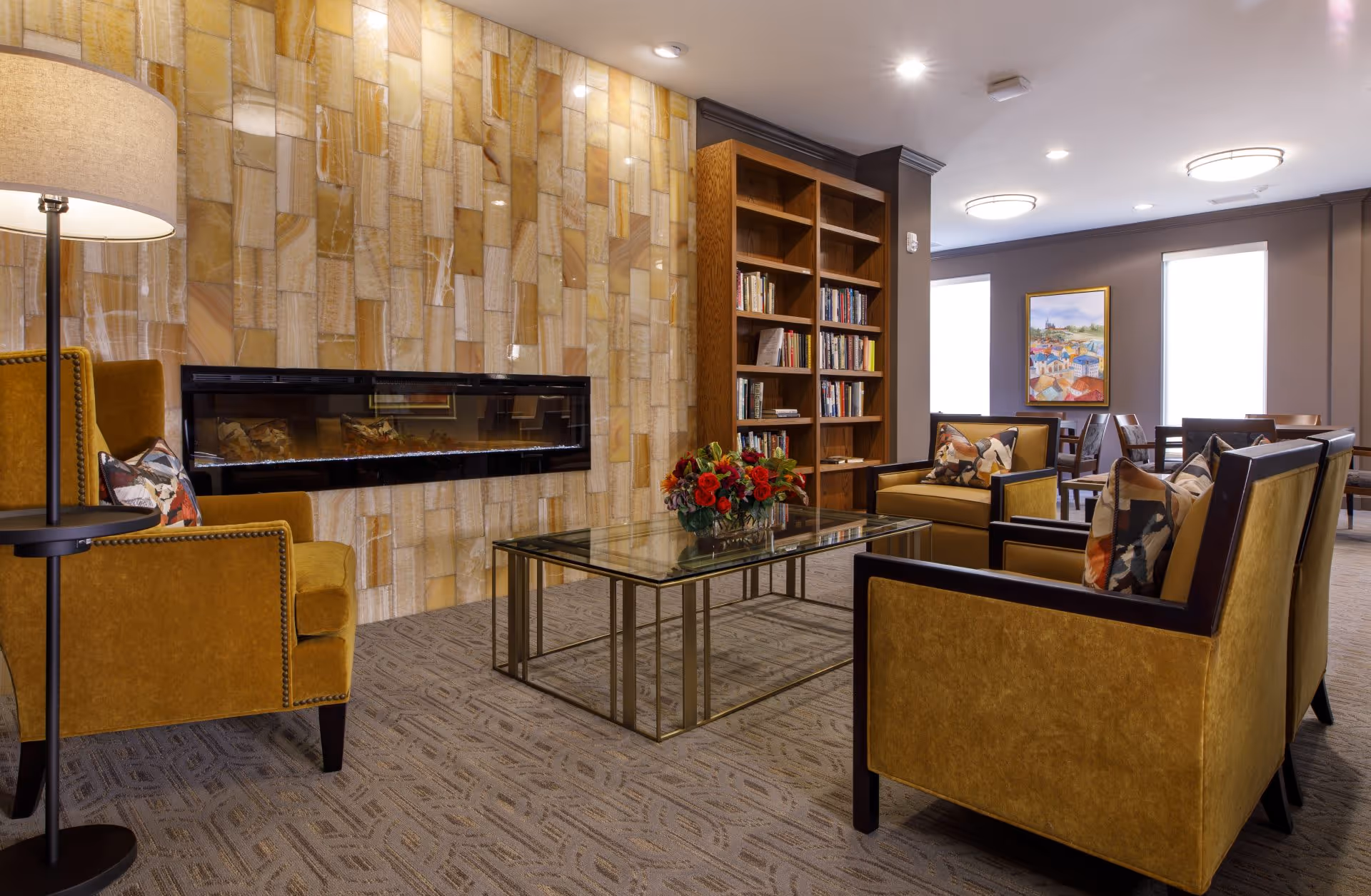 A cozy living room area with mustard yellow armchairs and a glass coffee table with a flower arrangement. The room features a modern electric fireplace set into a wall with a textured stone tile design, a wooden bookshelf filled with books, and a painting on the far wall between two large windows.