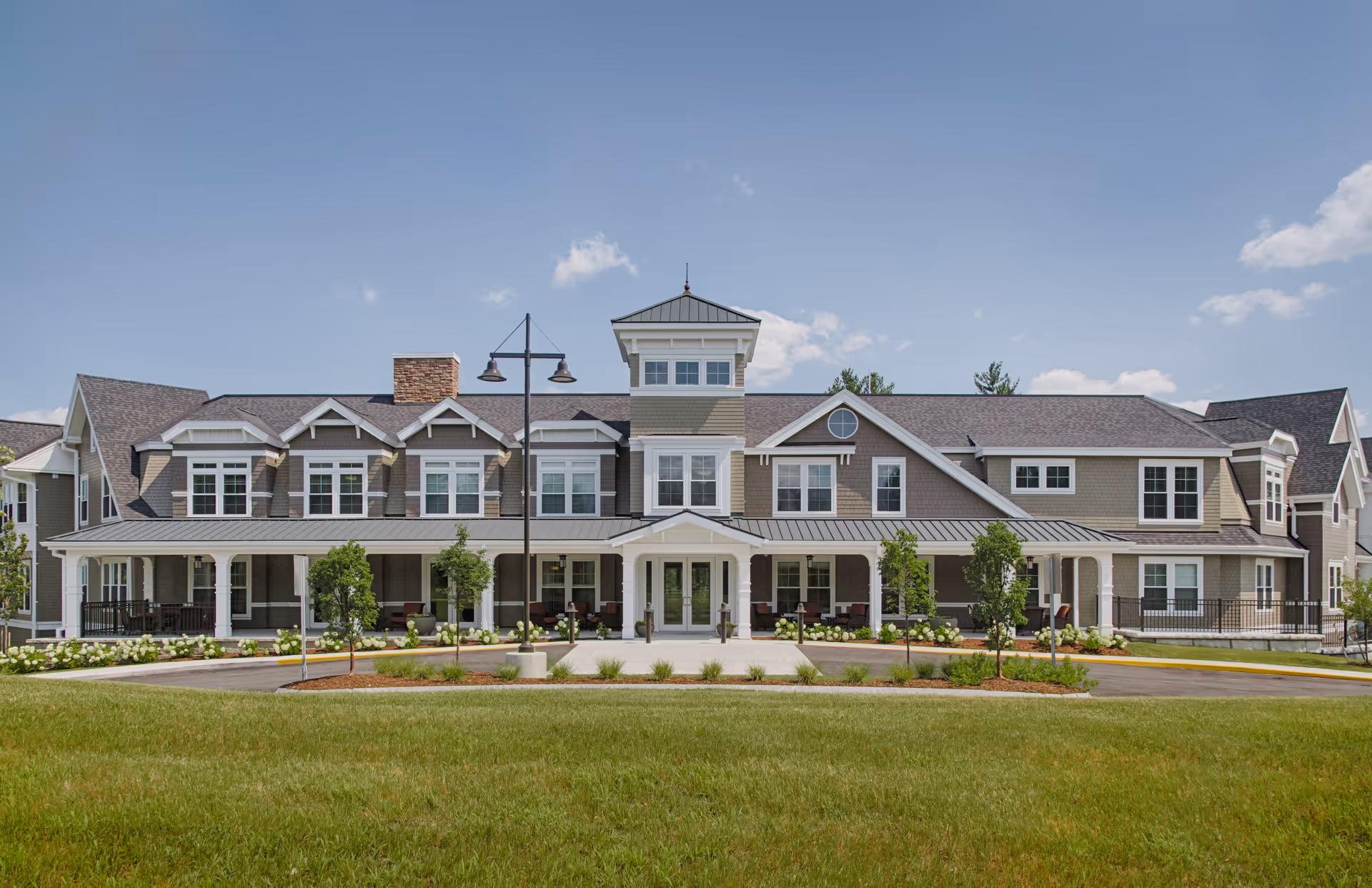 Front exterior view of The Residence at Salem Woods, a large two-story building with multiple windows, a covered entrance with white pillars, landscaped greenery, and a clear blue sky above.