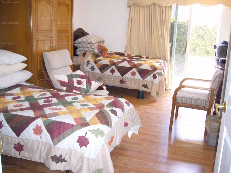 Two single beds with leaf-pattern quilts in a bright room with wood floors, a sliding glass door, wardrobe and chairs.