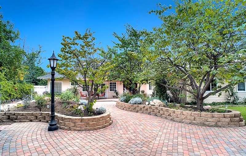 A paved courtyard with curved brick pathways and raised garden beds containing trees and shrubs. A black lamppost stands on the left side, and a single-story building with a porch and white doors is visible in the background under a clear blue sky.