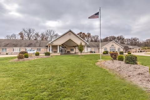 Front exterior view of Brookstone Estates of Paris, a single-story building with a covered entrance, an American flag on a flagpole in the foreground, and landscaped green lawn and bushes under a cloudy sky.