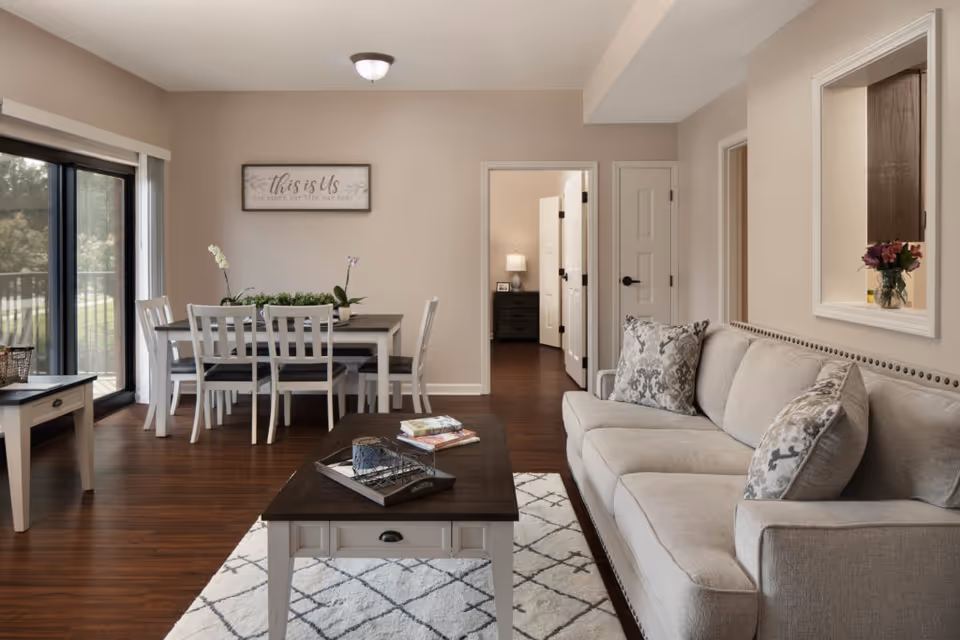 A cozy living room and dining area in a senior living facility. The living room features a beige sofa with patterned cushions, a dark wood coffee table on a light patterned rug, and a small side table with a basket. The dining area has a rectangular table with six white chairs and decorative plants on top. Large sliding glass doors let in natural light and offer a view of greenery outside. A framed sign on the wall reads 'this is Us'. In the background, an open doorway leads to another room with a lamp and nightstand.