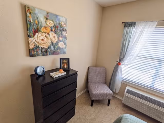 A corner of a bedroom featuring a black chest of drawers with a round clock, a small tray, and a framed photo on top. Above the chest hangs a floral painting. Next to the chest is a patterned upholstered chair near a window with sheer and solid curtains. Below the window is a wall-mounted air conditioning unit.