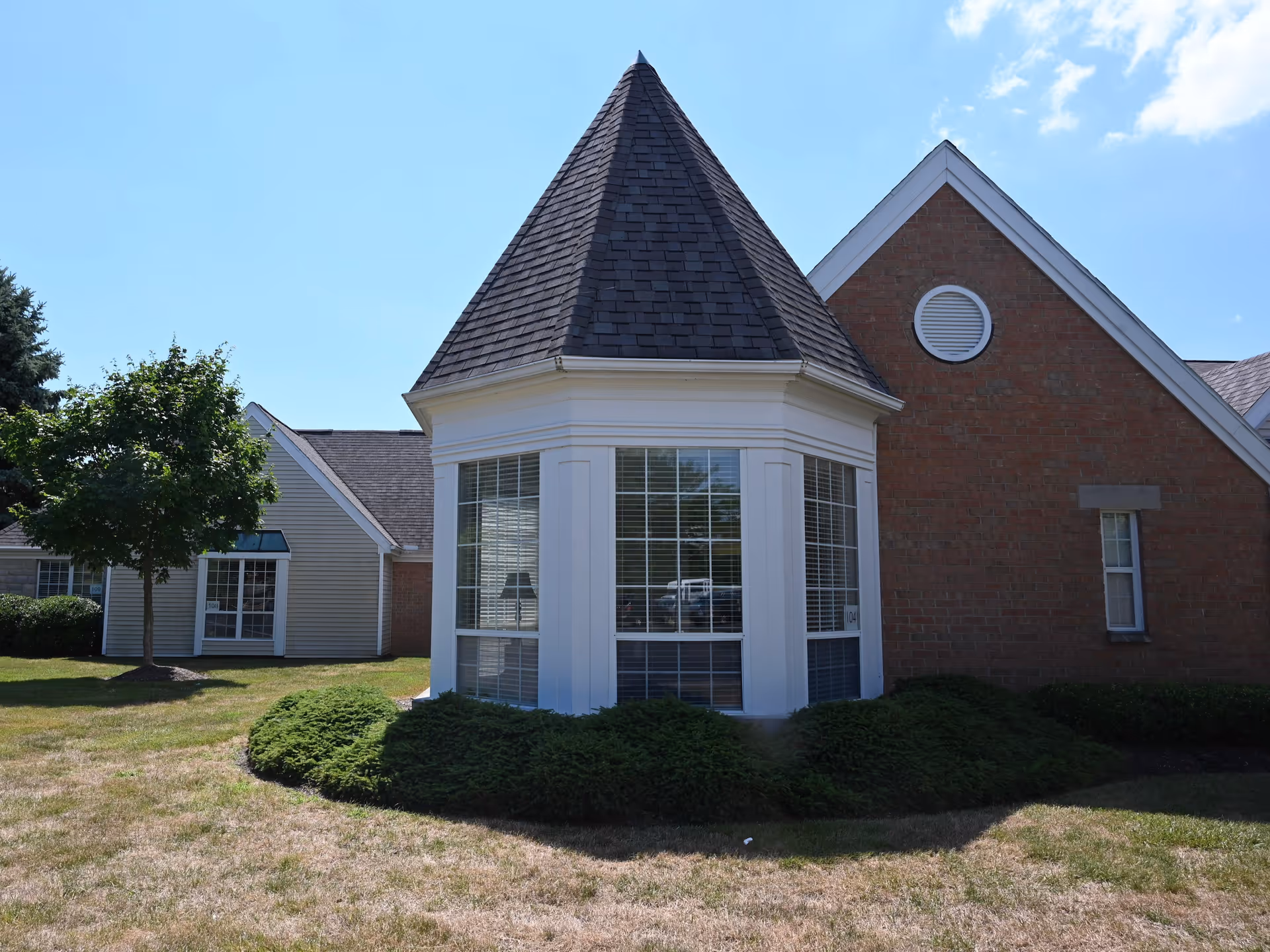Front exterior of a brick and siding building with a prominent octagonal bay window topped by a conical roof and surrounded by lawn and shrubs.