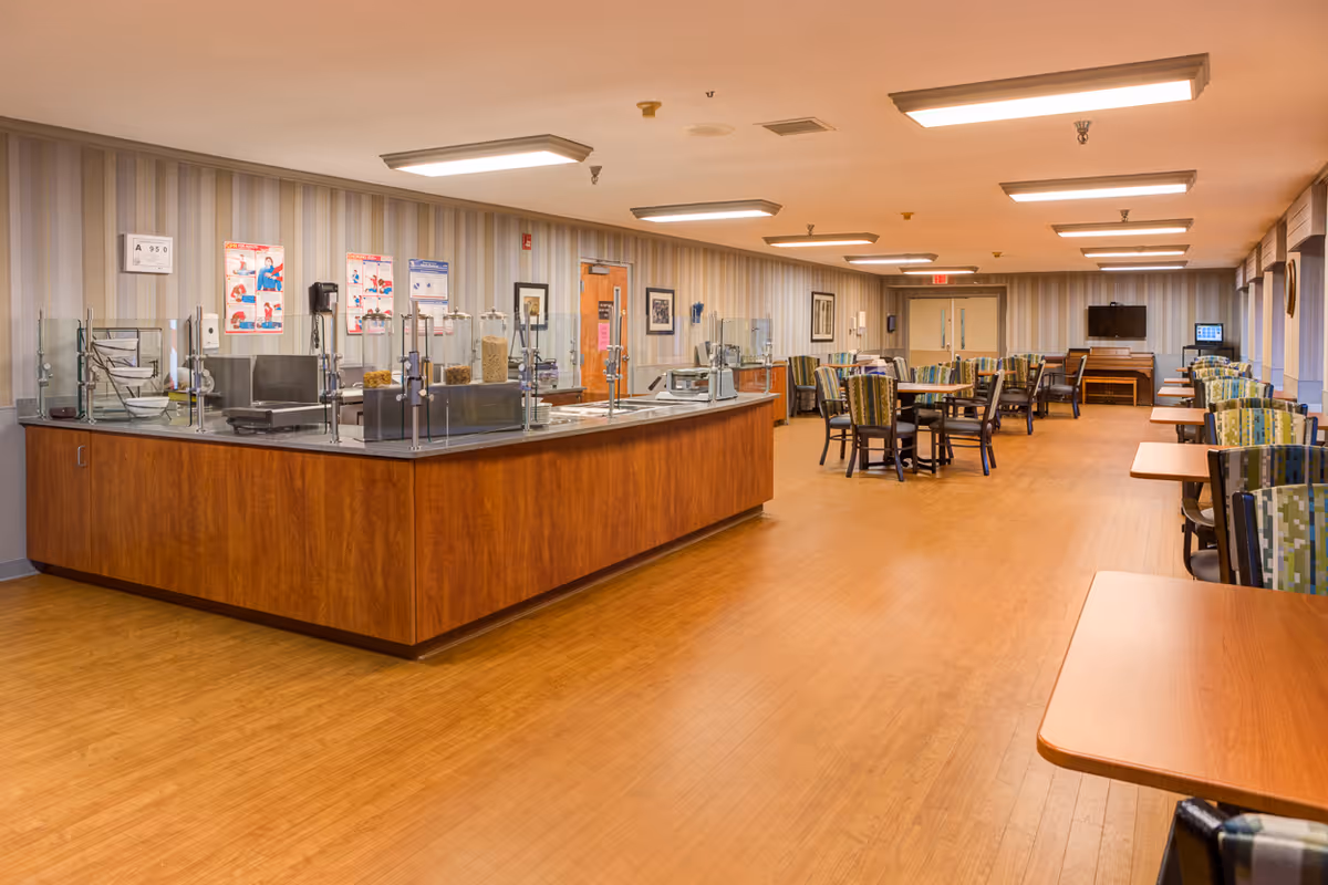 Spacious senior dining room with a central serving counter, multiple tables and chairs, and a TV at the far end.