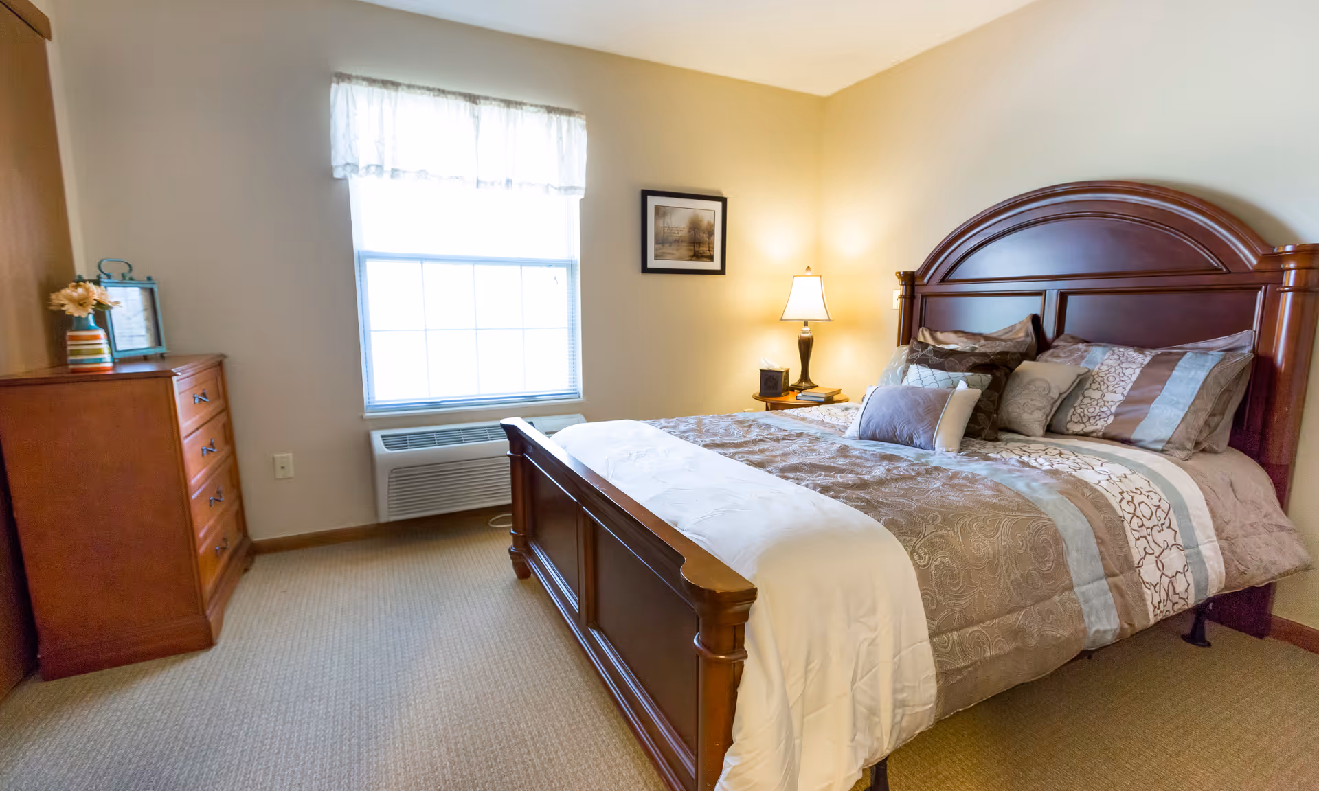 Well-lit bedroom with a large wooden bed dressed in patterned bedding, a nightstand with a lamp, and a dresser beneath a window.