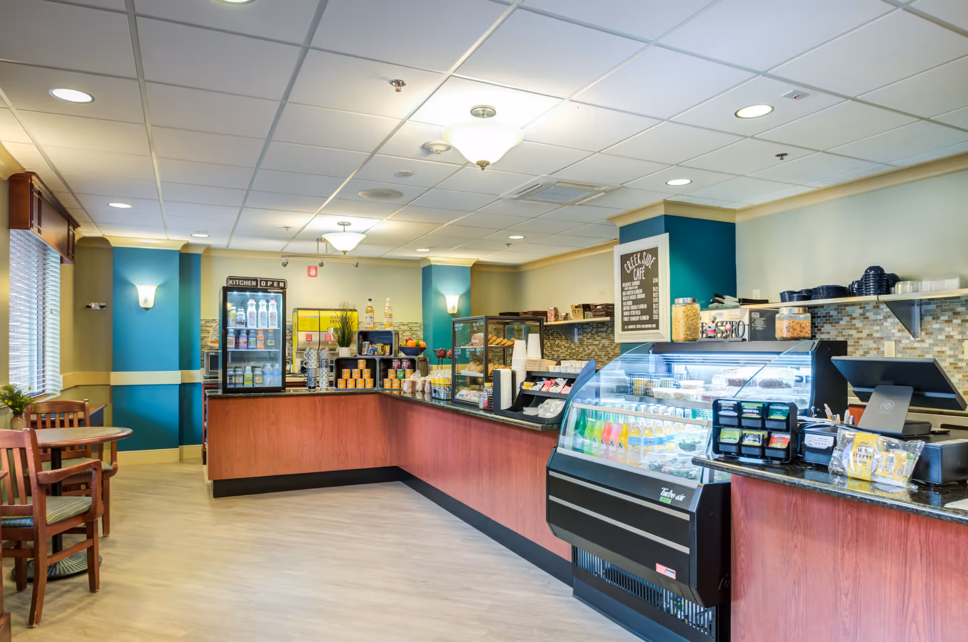 Interior view of a cafe area in a senior living facility with a counter displaying various snacks, beverages, and a refrigerated display case. There are wooden tables and chairs to the left, teal and beige walls, and ceiling lights providing bright illumination.