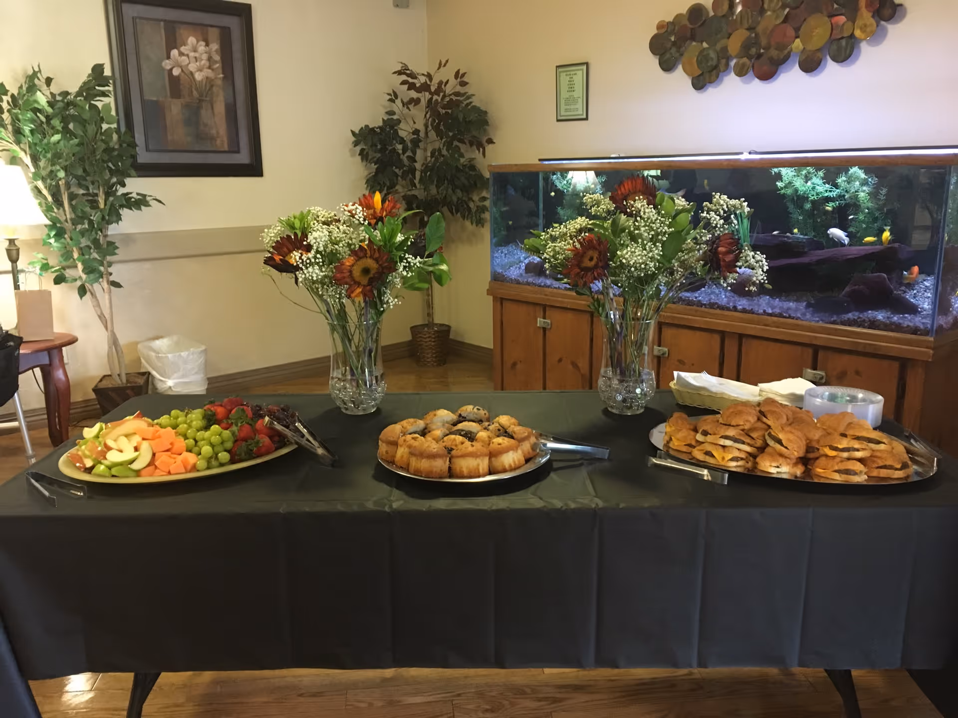 A buffet table covered with a black tablecloth displaying trays of fresh fruit, muffins, and sandwiches. Two vases with floral arrangements featuring sunflowers and baby's breath are placed on the table. In the background, there is a large fish tank with colorful fish, potted plants, a framed flower painting, and a decorative wall piece.
