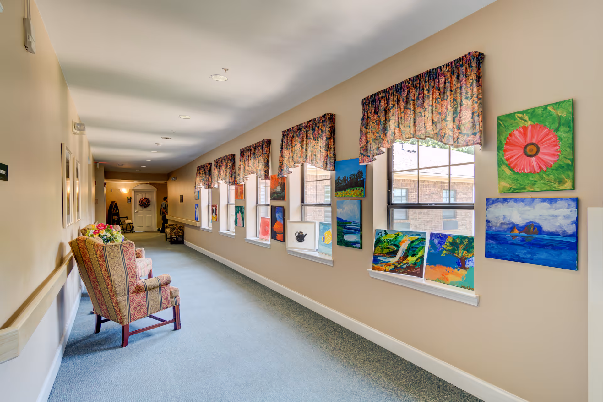 A long, well-lit hallway with beige walls and carpeted floor. Several colorful paintings are displayed on the wall beneath windows with floral valances. Two upholstered armchairs with floral patterns are placed along the left side of the hallway. The hallway leads to a white door at the far end.