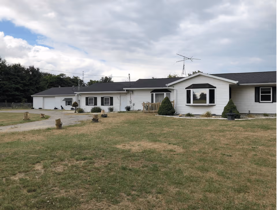 Single-story white building with black shutters and a large bay window, surrounded by a grassy lawn with some tree stumps and a paved driveway leading to a detached garage. The sky is partly cloudy.