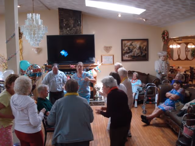 A group of elderly people and a caregiver gathered in a living room area decorated with balloons and a Happy Birthday banner. Some individuals are standing and interacting, while others are seated on couches and chairs. The room features a large flat-screen TV mounted above a fireplace, a chandelier, and wooden flooring.