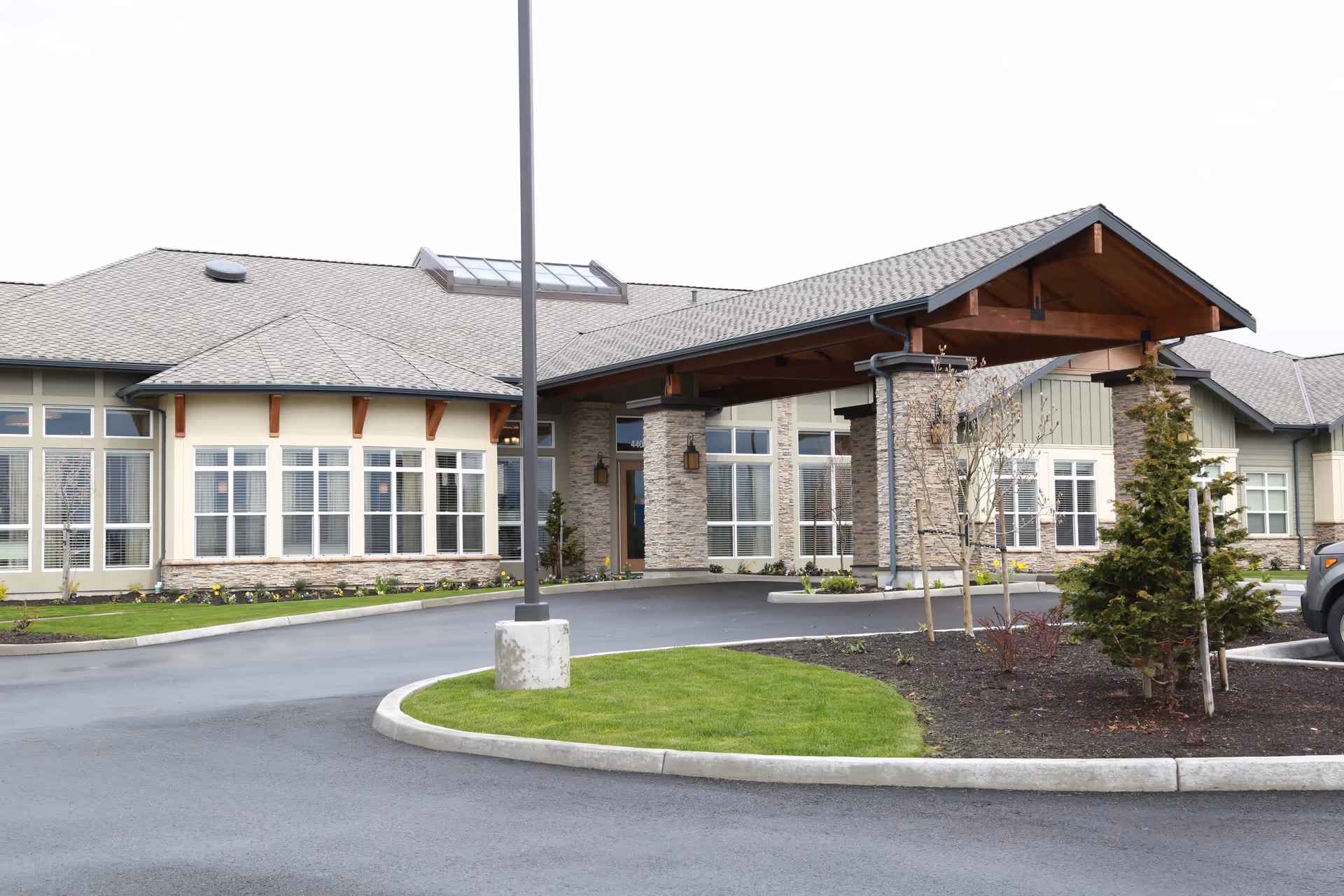 Exterior view of Silverado Bellingham Memory Care Community building with a covered entrance, stone pillars, large windows, and landscaped greenery including small trees and grass.