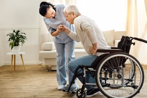 A caregiver is helping an elderly man in a wheelchair to stand up in a bright living room with wooden floors, a white sofa, and a potted plant on a small table.