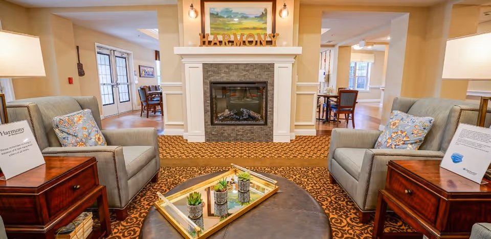 A cozy living room area in Harmony at Brentwood featuring two gray armchairs with floral pillows, a dark round ottoman with a gold tray holding small potted plants, and a fireplace with a stone surround and a painting above it. The word 'HARMONY' is displayed on the mantel. There are wooden side tables with lamps and informational signs on each side of the chairs. In the background, a dining area with tables and chairs is visible through open doorways.