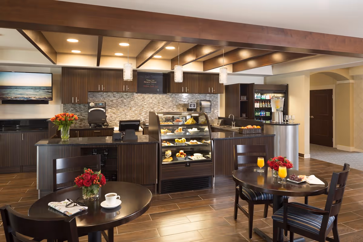 Interior view of a senior living facility cafe area with dark wood furniture and cabinetry. There are two round tables with chairs, each table decorated with a vase of red flowers. One table has a cup of coffee, newspaper, and glasses, while the other has two glasses of orange juice and a plate of snacks. Behind the tables is a counter with a glass display case containing pastries and fruit. The back wall features a mosaic tile backsplash, cabinets, a coffee machine, and a refrigerator stocked with beverages. Pendant lights hang from the ceiling with wooden beams.