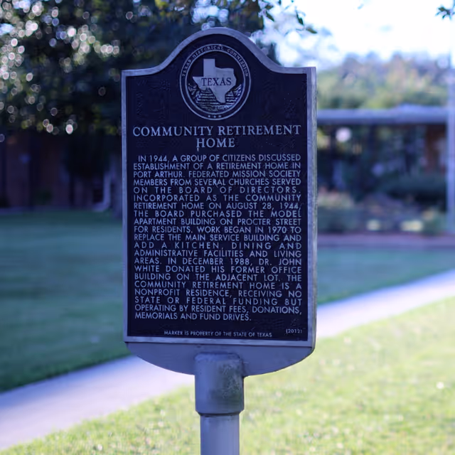 A historical marker sign for the Community Retirement Home in Texas, detailing its establishment in 1944, development history, and nonprofit status. The sign is mounted on a post with a blurred background of grass, trees, and a pathway.