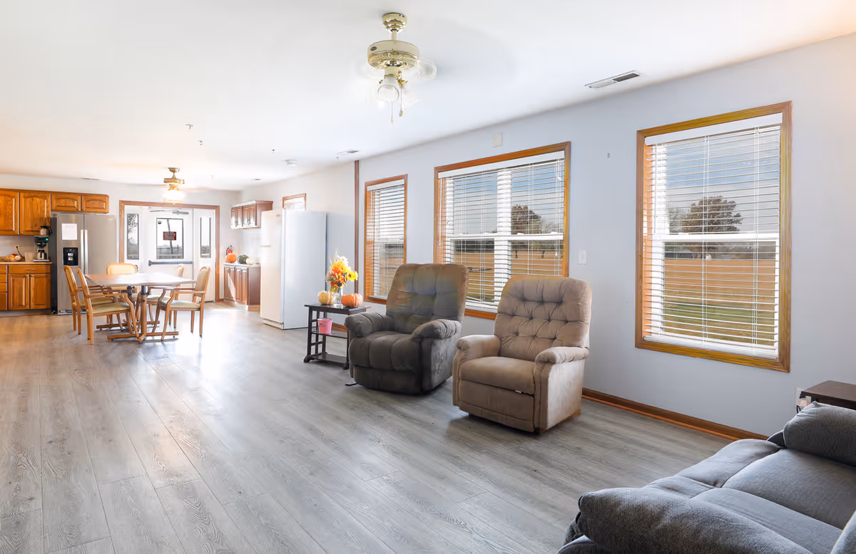 A bright and spacious common area in a senior living facility featuring two recliner chairs and a sofa near three large windows with blinds. The room has light-colored walls and wood flooring. In the background, there is a dining area with a table and chairs, wooden kitchen cabinets, a refrigerator, and a door leading outside.