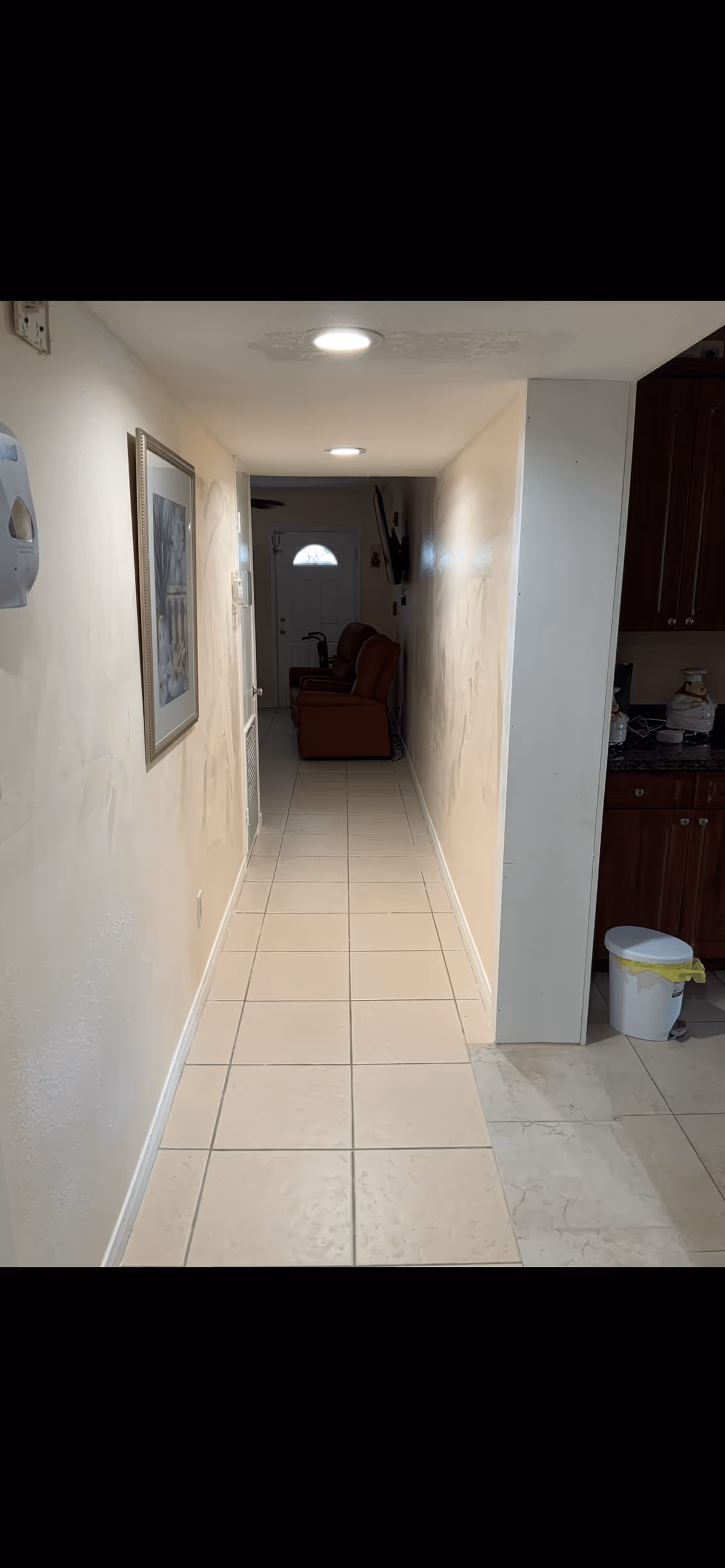 A narrow hallway with beige tiled floor and cream-colored walls. On the left wall, there is a framed picture and a wall-mounted dispenser. At the end of the hallway, two brown recliner chairs are positioned facing a wall-mounted TV. To the right, part of a kitchen area with dark wooden cabinets and a white trash bin with a yellow liner is visible.