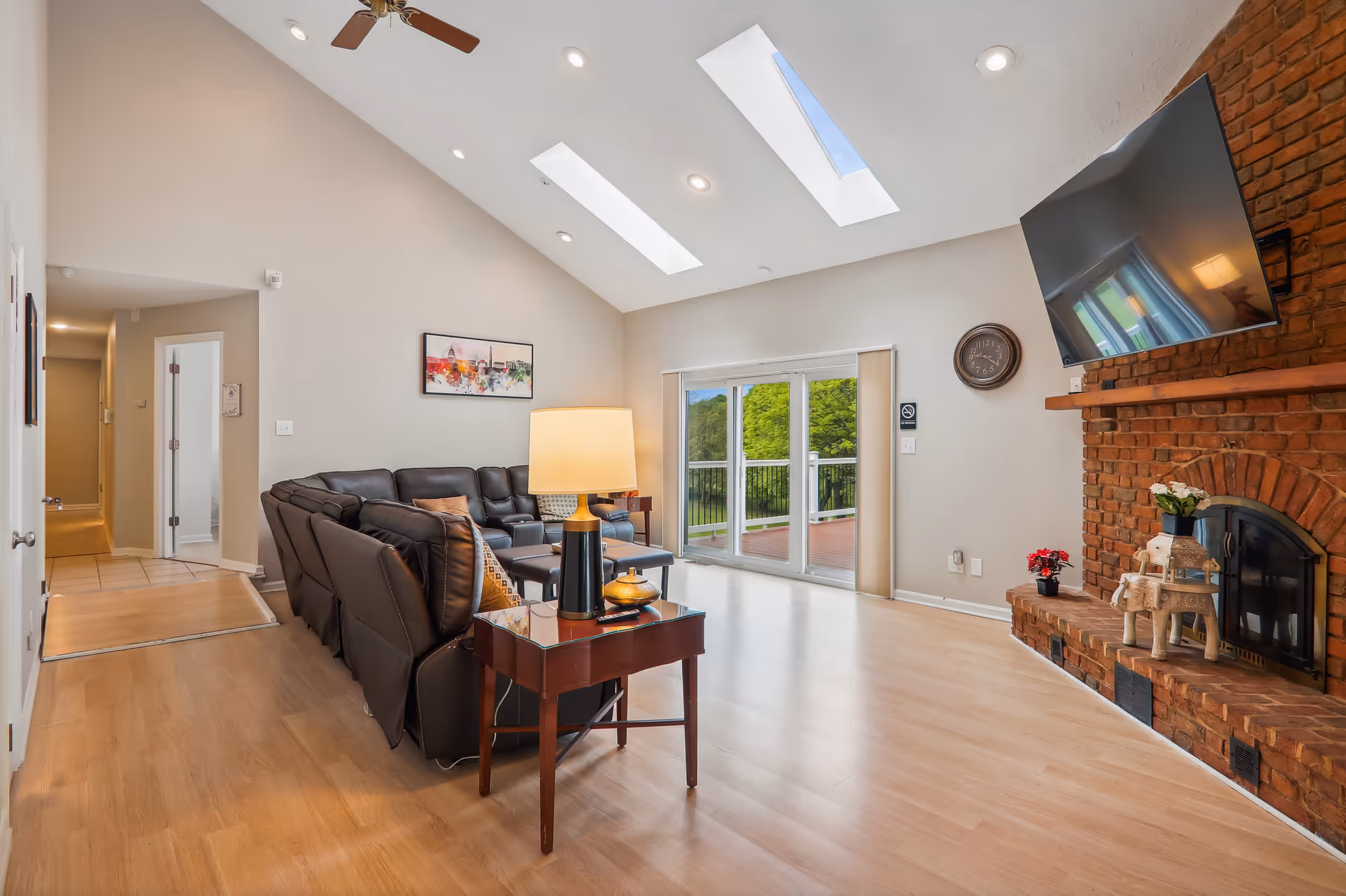 A bright living room with light wood flooring, a dark brown leather sectional sofa, a wooden side table with a lamp, and a brick fireplace with a mounted flat-screen TV above it. The room has two skylights in the vaulted ceiling and large sliding glass doors leading to an outdoor deck with a view of green trees.