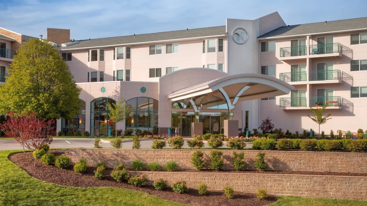Front entrance of a multi-story senior living building with a covered porte-cochère, balconies, and landscaped terraces.