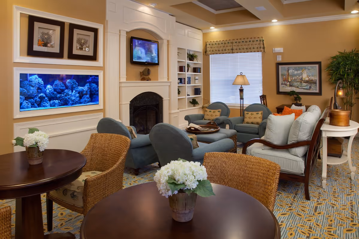 A cozy living room with beige walls and patterned carpet. The room features a built-in fireplace with a TV mounted above it, a large aquarium built into the wall, and built-in shelves with decorative items. There are several upholstered chairs and sofas arranged around a coffee table, wicker chairs around small round tables with flower arrangements, a floor lamp near a window with blinds, and framed artwork on the walls.
