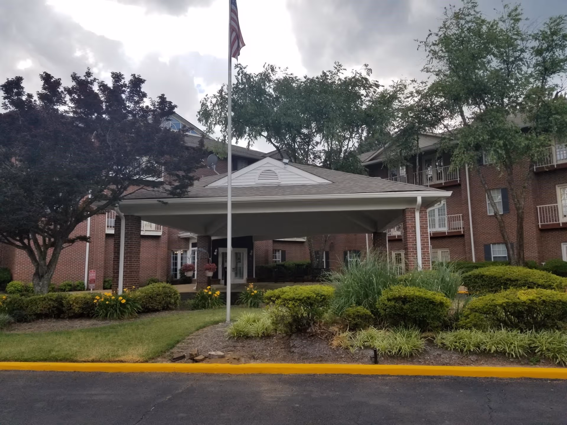 Front exterior view of a senior living facility named Kennington Pointe with a covered entrance, an American flag on a flagpole, well-maintained landscaping including bushes, trees, and flowers, and a brick building with balconies and windows in the background under a cloudy sky.
