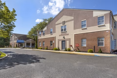 Exterior view of a two-story senior living facility building with a brick and beige facade under a partly cloudy sky. There is a paved driveway in front and some greenery along the building.
