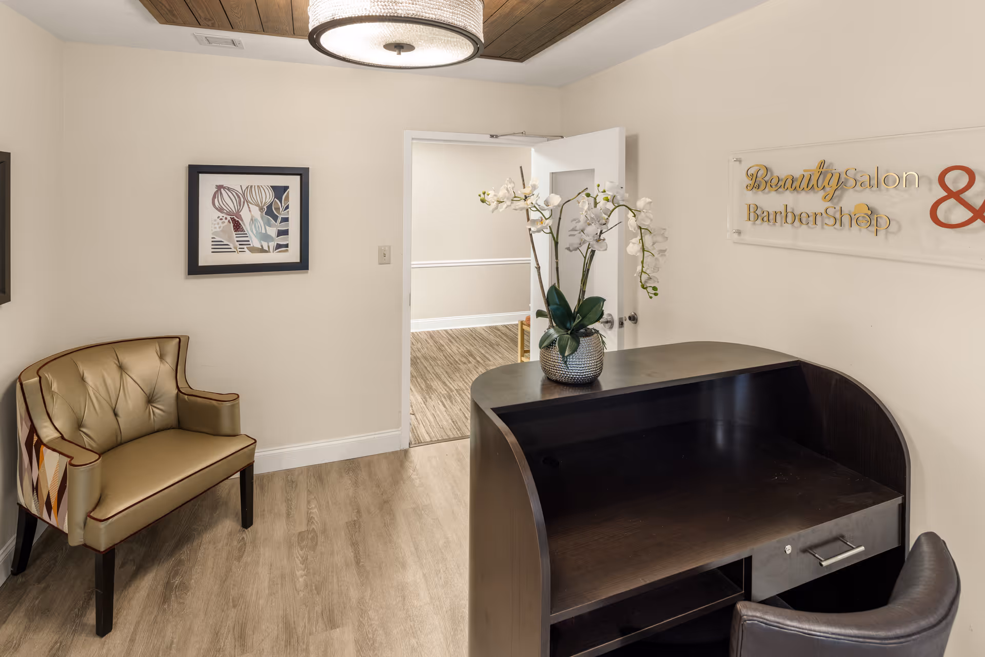 Reception area with a dark wooden desk and a black chair. A vase with white orchids is placed on the desk. To the left, there is a cushioned chair with a patterned backrest. A framed artwork hangs on the wall. The door is open, showing a hallway with wood flooring. A sign on the wall reads 'Beauty Salon & Barber Shop'.
