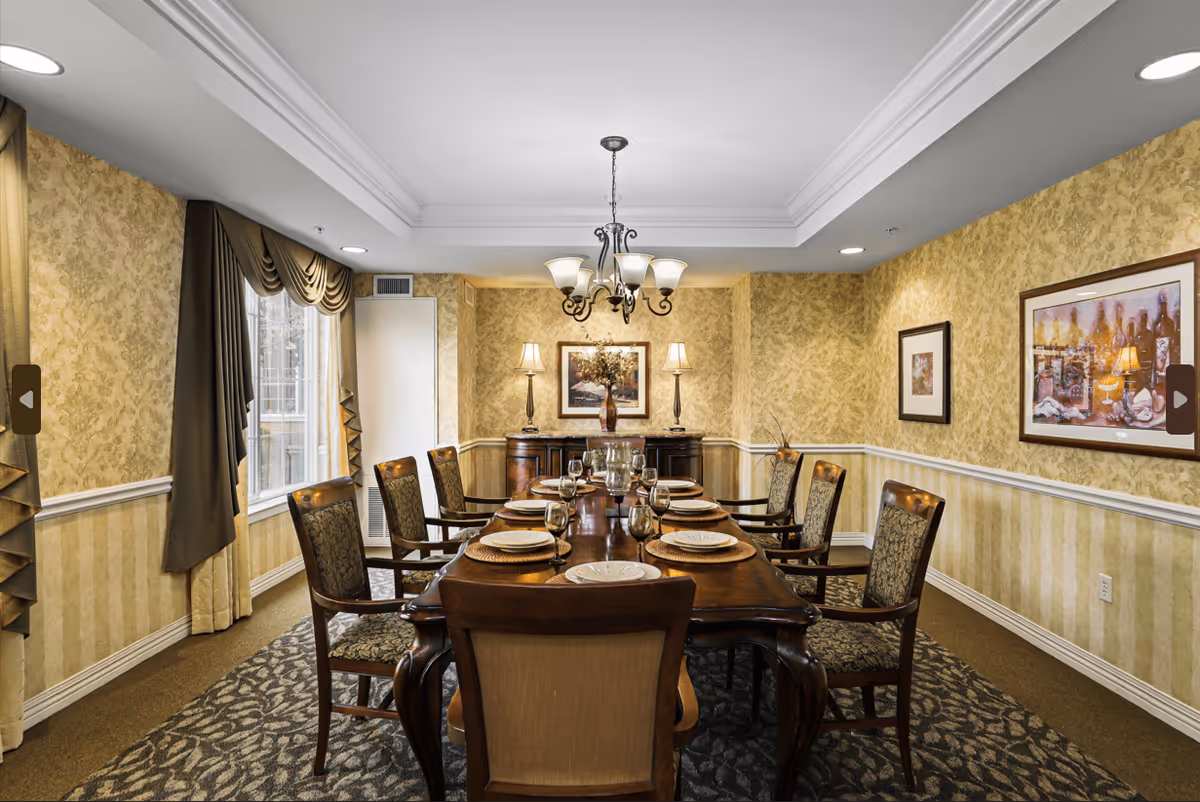 A formal dining room with a long wooden table set for eight people with plates, glasses, and napkins. The room has patterned wallpaper, a chandelier hanging from the ceiling, two table lamps on a sideboard, framed artwork on the walls, and large windows with draped curtains.