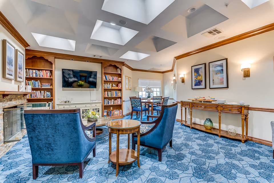 A cozy living room area with blue upholstered chairs, a round wooden coffee table, and a fireplace with a stone surround. The room features built-in wooden bookshelves filled with books, a wall-mounted flat-screen TV, and a wooden console table with decorative items. The ceiling has multiple skylights allowing natural light to brighten the space, and the floor is covered with a blue patterned carpet. There are framed artworks on the walls and a small dining table with chairs near a window.