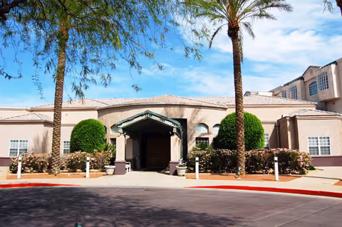 Exterior view of a senior living facility building with a covered entrance, palm trees, trimmed bushes, and clear blue sky.
