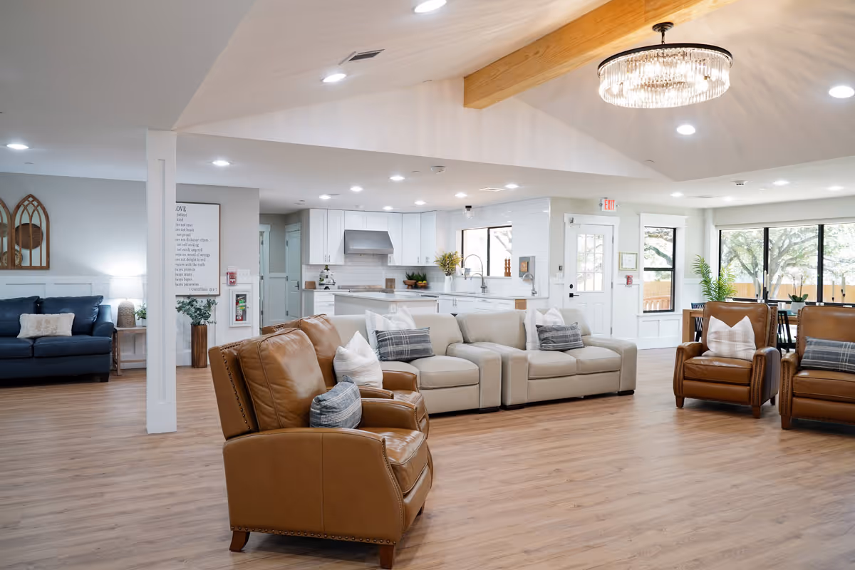 Open, well-lit assisted living common area with leather and fabric sofas and armchairs facing an adjacent kitchen and dining area.