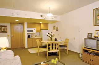 Interior view of a senior living facility room featuring a small dining area with a round wooden table and four chairs. The room includes a kitchenette with cabinets, a microwave, and a coffee maker. There is a wooden TV stand with an older-style television and framed pictures on the wall. A couch and a lamp are partially visible in the foreground.