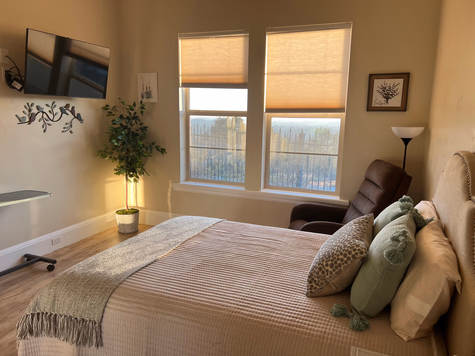 A cozy bedroom with a neatly made bed featuring multiple pillows and a textured blanket. There are two large windows with blinds partially drawn, allowing natural light to fill the room. A brown recliner chair and a floor lamp are positioned near the windows. A flat-screen TV is mounted on the wall opposite the bed, and a potted plant sits in the corner next to the windows. The room has light-colored walls and wooden flooring.