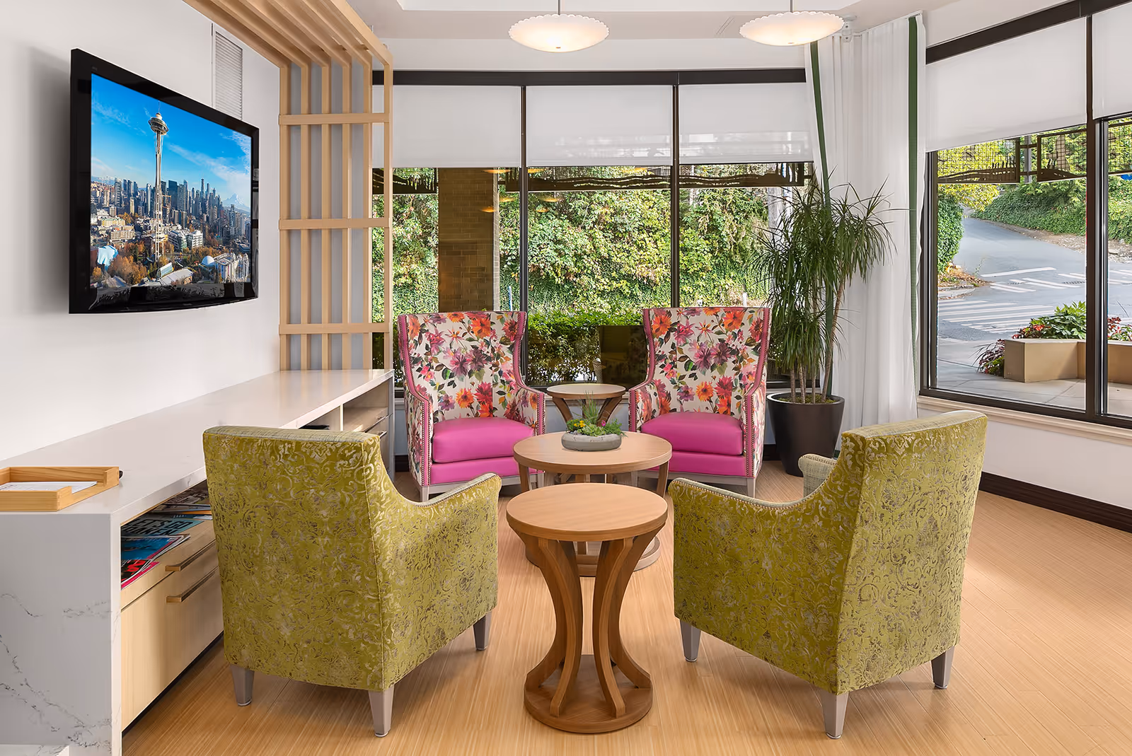 A bright and modern sitting area with two floral-patterned armchairs with pink cushions and two green armchairs arranged around three small wooden tables. A flat-screen TV mounted on the wall displays an image of the Seattle skyline with the Space Needle. Large windows provide a view of greenery and a driveway outside. A tall potted plant is placed near the windows.