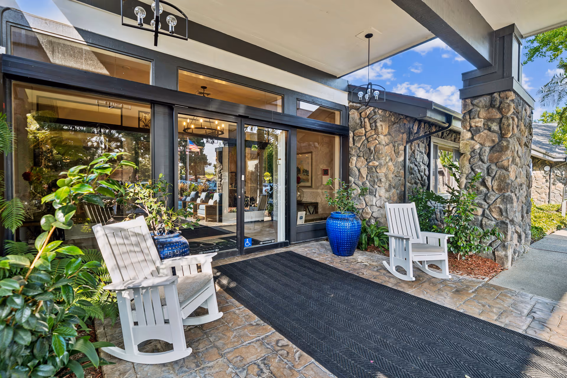Entrance to a senior living facility with stone pillars and walls, two white rocking chairs on a stone patio, large blue ceramic pots with green plants, and glass double doors leading inside. The interior visible through the doors shows a well-lit lobby area with seating and decorative lighting fixtures.