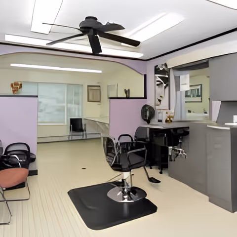 Interior view of a hair salon area with styling chairs, mirrors, a ceiling fan, and a reception counter. The walls are painted light purple and white, and the floor is light-colored. There are additional chairs and a window in the background.