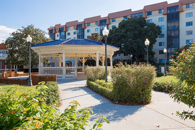Outdoor garden area with a white gazebo featuring a blue roof, surrounded by green bushes and flowering plants. There are several lamp posts along a paved walkway, and a multi-story residential building is visible in the background under a clear blue sky.
