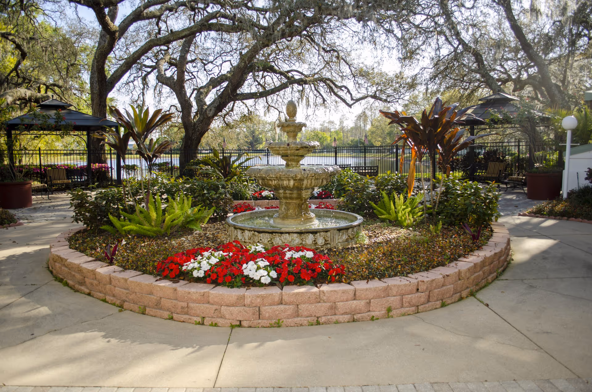 A landscaped outdoor garden area featuring a tiered stone water fountain surrounded by colorful flowers and green plants. There are large trees providing shade, black metal fencing in the background, and two gazebo structures with seating underneath. The scene overlooks a body of water with trees beyond.