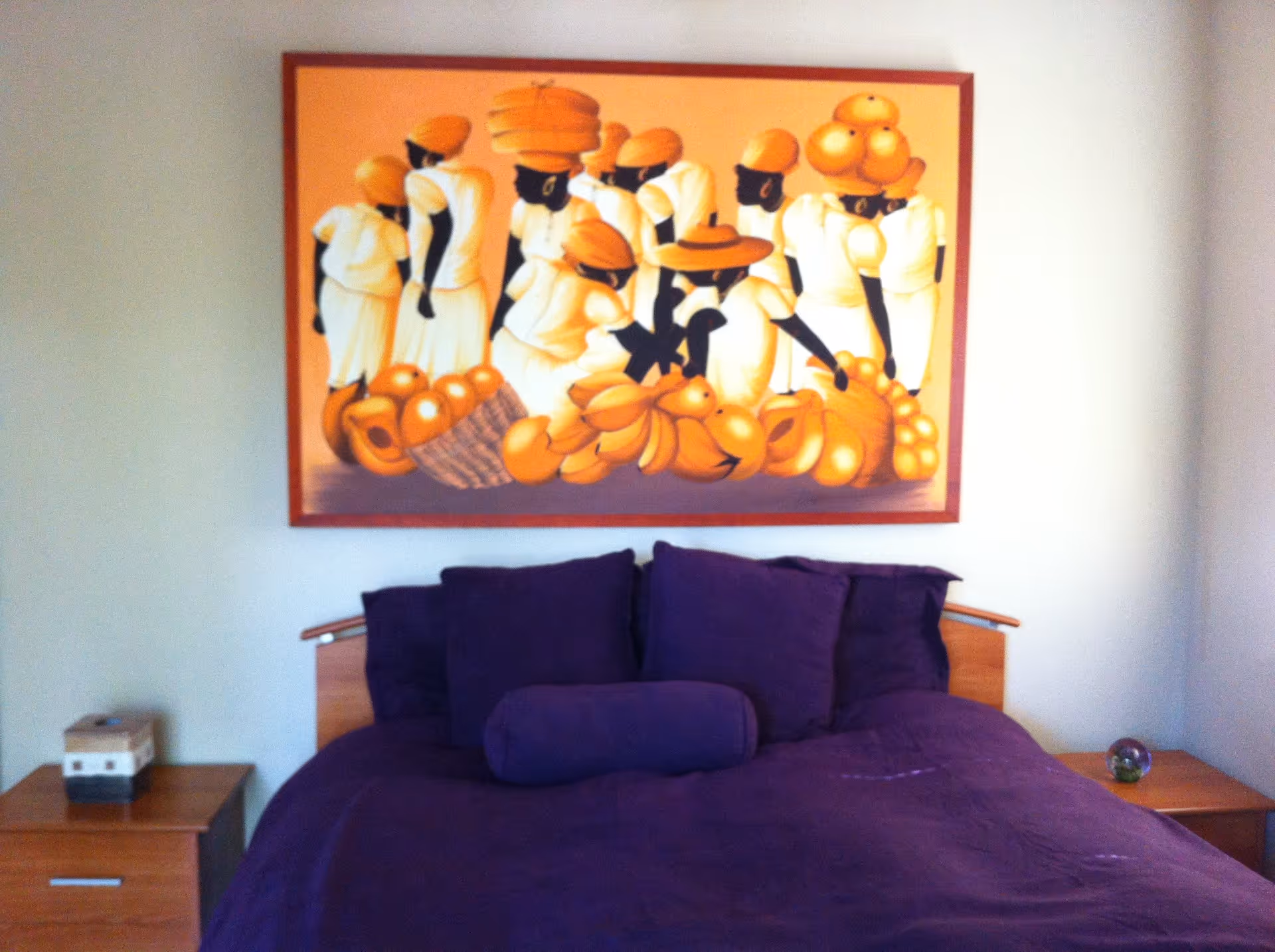 A bedroom with a bed covered in purple bedding and pillows. Above the bed hangs a large colorful painting depicting people in white clothing and orange headwear carrying baskets of fruit. There are wooden nightstands on either side of the bed, one with a small box and the other with a decorative glass orb.