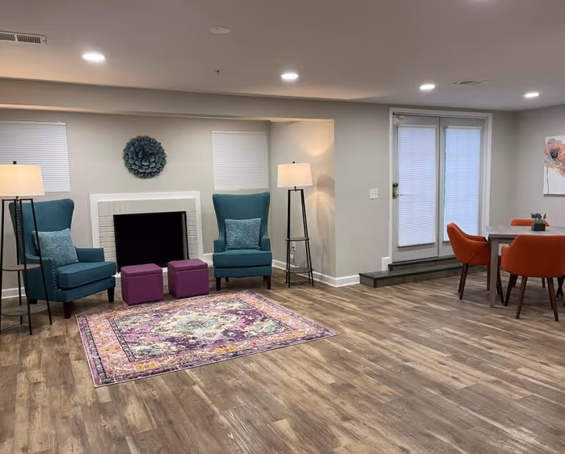 A cozy living room area with two teal armchairs flanking a white fireplace, two purple ottomans in front of the fireplace, a colorful patterned rug on wooden flooring, two tall floor lamps, and a dining table with four orange chairs near glass doors with blinds.