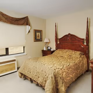 A furnished bedroom with a wooden four-poster bed covered in a patterned gold bedspread, a nightstand with a lamp, window with a blind, and framed wall art.