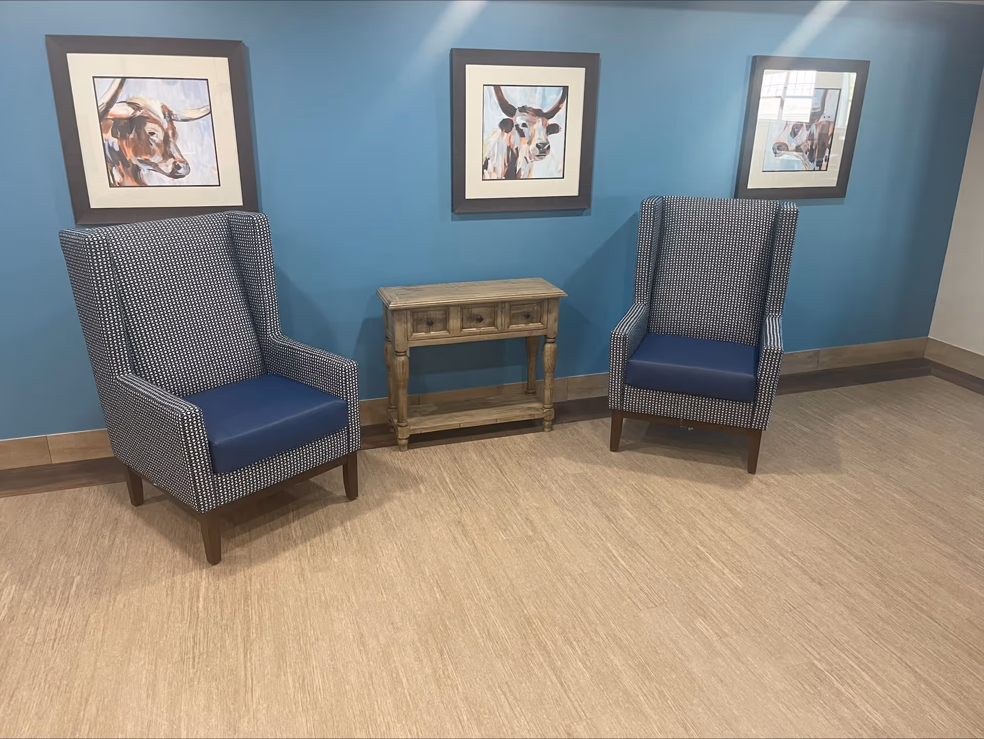 Two patterned armchairs with blue cushions are placed on either side of a small wooden table against a blue wall. Above the table, three framed paintings of cows are hung on the wall. The floor is light wood or laminate.